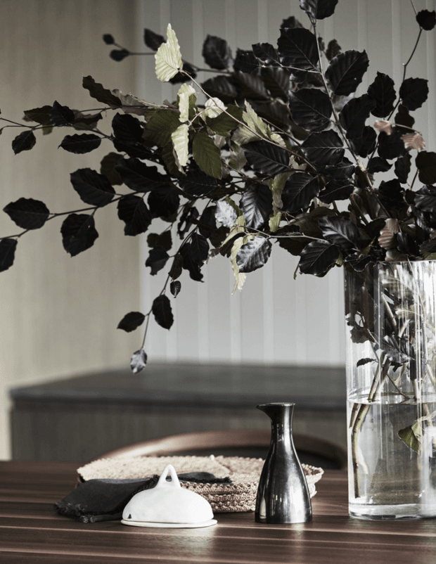 Styled interior vignette featuring a dark timber dining table with a white ceramic object, black metal vessel, folded dark textile, and a tall clear glass vase filled with dramatic dark leafy branches. The background includes subtle vertical wall panelling and a built-in upholstered bench, creating a moody, refined decorative arrangement.