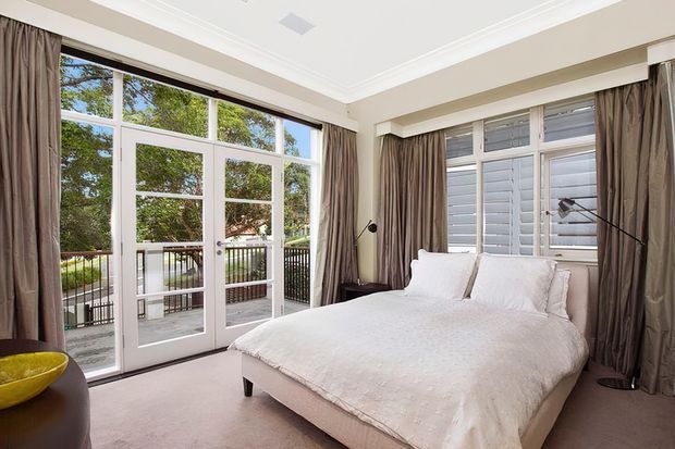 Bright, elegant bedroom with a large upholstered bed dressed in white linens, full-height glazed French doors opening to a balcony, and additional tall windows with exterior shutters. Taupe floor-to-ceiling curtains, pale carpet, white decorative ceiling cornice, and a simple floor lamp create a refined, serene interior.