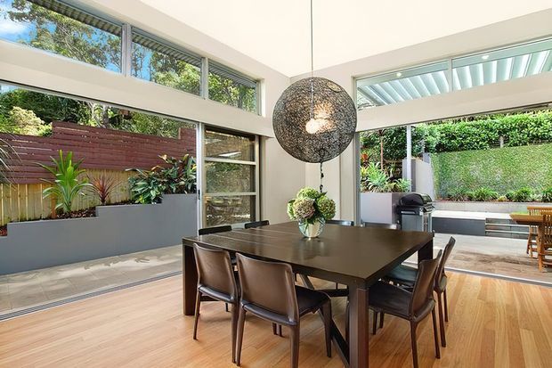 Bright contemporary dining area with a dark rectangular dining table and six leather-upholstered chairs, opening onto a landscaped courtyard through extensive glazing. The room features warm timber flooring, pale walls, high-level windows, a large sculptural woven pendant light, and an outdoor barbecue area visible beyond.