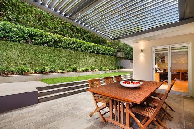 A landscaped courtyard terrace with a timber outdoor dining table and folding chairs beneath a translucent louvred pergola. Stepped garden beds, a lush planted wall, paved flooring, and a glazed doorway into the interior create a sheltered indoor-outdoor entertaining space.