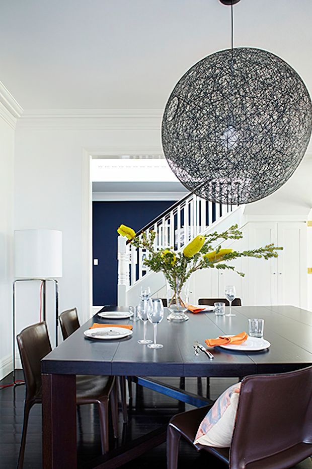 A bright, refined dining room with a large dark rectangular dining table, leather dining chairs, white cabinetry, and a dramatic spherical woven pendant light. A staircase with white balustrades and a dark navy wall are visible beyond the dining area. The room combines crisp white architectural detailing with dark timber or stained-wood furniture and restrained contemporary styling.