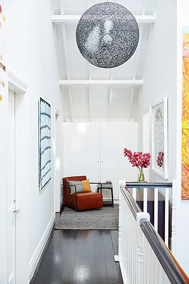 Bright, narrow upper-level hallway or landing with dark timber flooring, white walls, detailed trim, and a white painted exposed-beam ceiling. The corridor opens into a small seating nook with a rust-red armchair, side table, grey rug, framed artwork, and a vase of pink flowers. A large textured spherical pendant hangs overhead, while a white balustrade with dark timber handrail lines the right side.