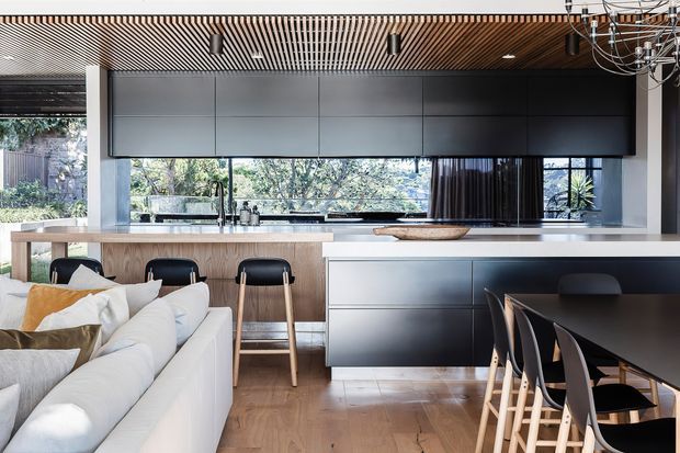 Open-plan contemporary kitchen and dining area viewed from the living zone, featuring a large island with timber stools, dark grey built-in cabinetry, a pale benchtop, timber flooring, a black dining table with bar-height chairs, and a slatted timber ceiling. Large windows provide views to greenery outside.