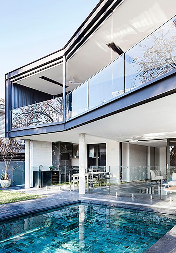 Modern multi-level residence with strong horizontal lines, white rendered surfaces, dark metal framing, extensive glass balustrades and open-plan entertaining areas overlooking a tiled swimming pool. The upper level includes a covered balcony with outdoor seating, while the ground level features a partially enclosed patio and dining area.