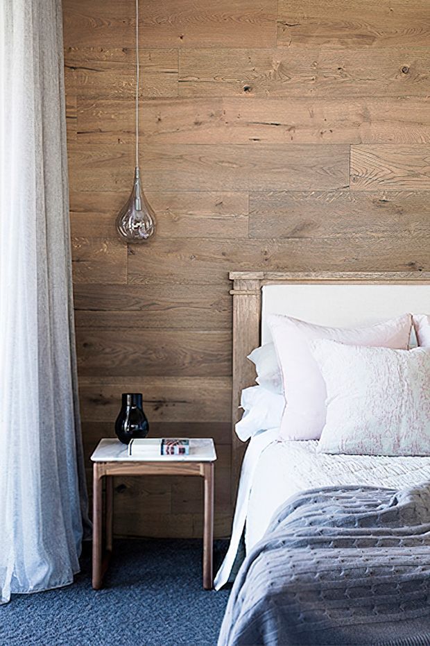 A softly styled bedroom vignette featuring a partially visible bed with white and pale pink bedding, a grey textured throw, a warm reclaimed timber feature wall, and matching timber bedside table. A black vase, small book, exposed pendant light, sheer grey curtains, and dark carpet complete the calm, rustic-contemporary interior.