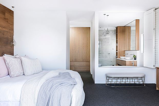 Bright contemporary bedroom with a white upholstered bed, pale bedding, and a timber feature wall. A large mirror reflects an adjacent bathroom with a timber vanity, wall-mounted basin, freestanding bathtub, and glass shower enclosure. Dark carpet contrasts with the light walls and timber joinery.