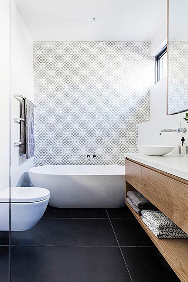 A bright, compact contemporary bathroom featuring a freestanding white bathtub, wall-mounted toilet, floating timber vanity with open shelving, vessel basin, and wall-mounted tapware. The walls are finished in a small white geometric-patterned tile, contrasted by large matte black floor tiles. A tall mirror and high window bring additional light into the room.