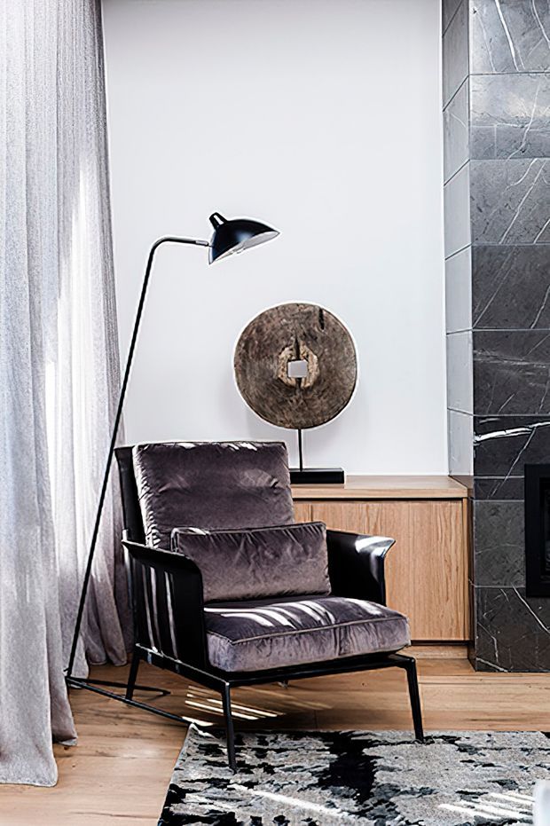 A refined living room corner featuring a black-framed upholstered lounge chair with dark velvet cushions, a slim black floor lamp, and a round sculptural wall light or artwork above a timber sideboard. The space has pale walls, timber flooring, sheer curtains, a dark patterned rug, and a dramatic dark stone-tiled fireplace surround at the right edge. The styling is contemporary with a moody, sophisticated palette.