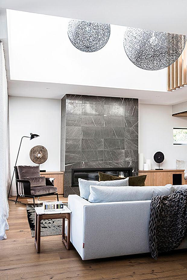 Bright contemporary living room with a large pale sectional sofa, timber flooring, a dark stone-tiled fireplace feature wall, integrated timber cabinetry, sculptural pendant lights, and an armchair with floor lamp. A partial stair balustrade and high ceiling add architectural interest.