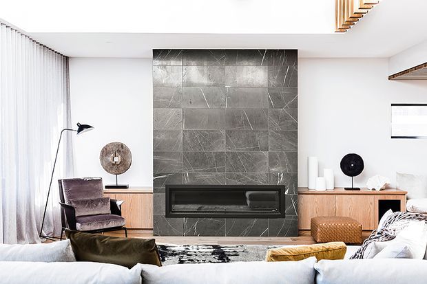Bright contemporary living room featuring a large dark stone-tiled fireplace wall with a built-in linear firebox, low timber cabinetry, pale upholstered sofas, accent chairs, and floor-to-ceiling sheer curtains. Timber ceiling detailing and carefully styled decorative objects add warmth and texture.