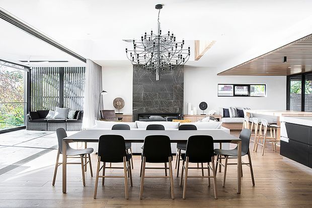 Bright, open-plan contemporary living space combining a dining area, living room and kitchen. A long white dining table with mixed black and grey chairs sits on a light timber floor. The living area includes a dark sofa and a textured dark feature wall with a fireplace or low media unit, while the kitchen has a dark island with timber accents and bar stools. Large glazing, vertical blinds, a timber-lined ceiling section and a sculptural multi-arm chandelier add architectural interest.