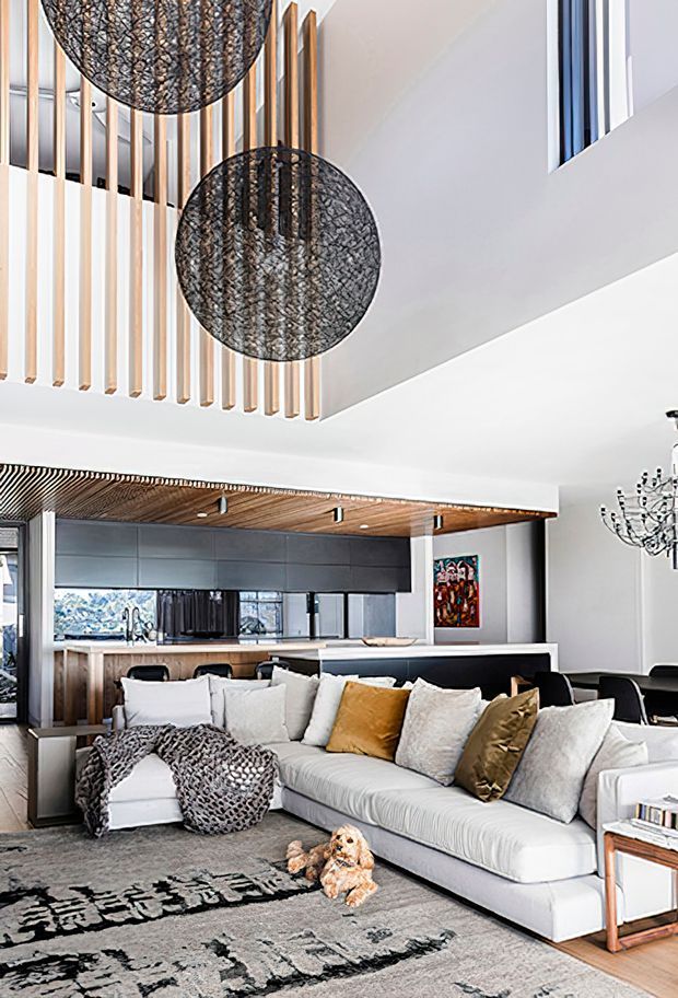 Bright open-plan contemporary living space with a large pale sectional sofa, patterned rug, layered neutral and ochre cushions, and sculptural dark pendant lights. The room opens toward a modern kitchen and dining area, with timber ceiling slats, vertical timber screening, dark cabinetry, white walls, and a tall void or high ceiling. A chandelier and colourful artwork add decorative accents.