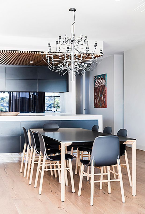 A bright contemporary open-plan dining area with a large rectangular timber dining table and dark upholstered chairs, positioned beside a sleek dark kitchen. The space features pale timber flooring, white walls, dark built-in cabinetry, a striking multi-arm chrome chandelier, and a slatted timber ceiling detail over the kitchen.
