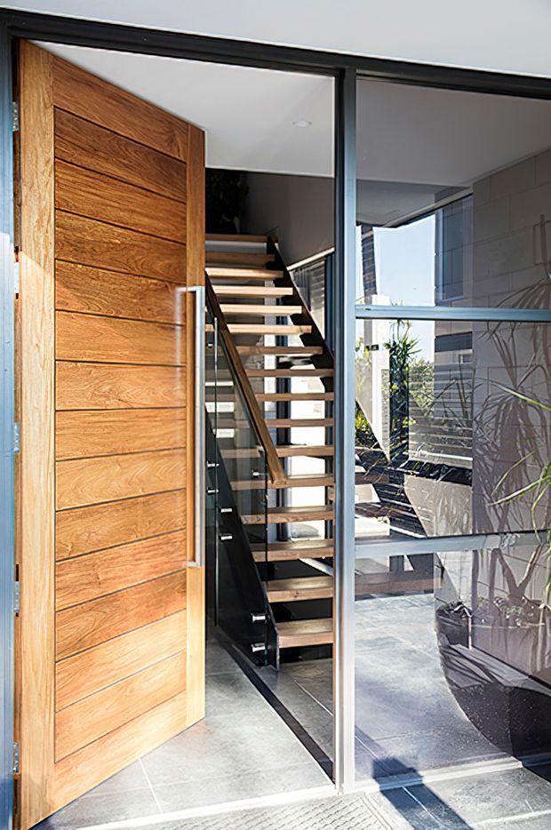 Contemporary residential entry viewed through an open pivot door, featuring a warm horizontal-grain timber door, dark metal-framed glazing, and a modern floating staircase with timber treads and glass balustrade. A tiled entry floor and bright internal courtyard or living area are visible beyond.