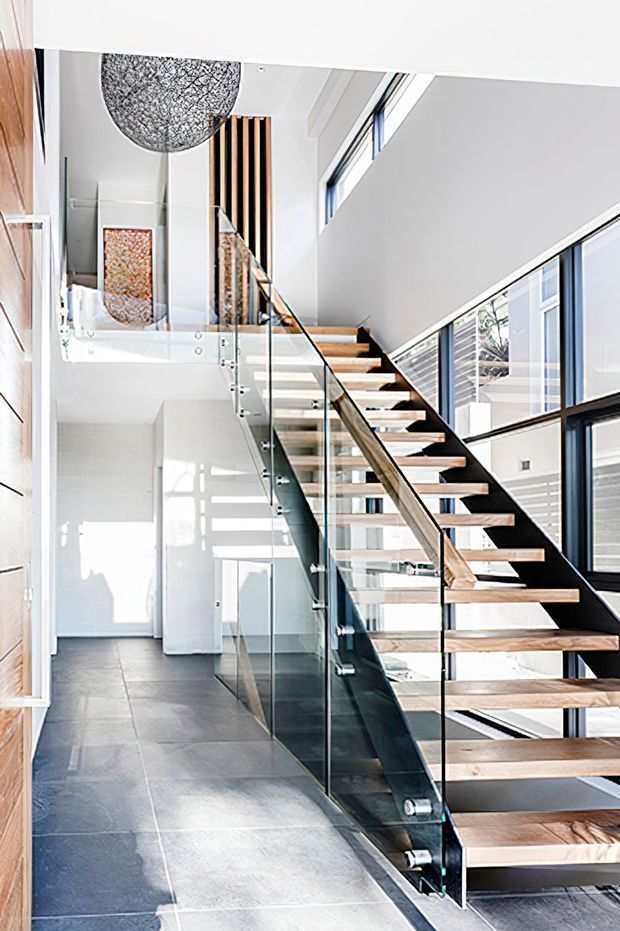 Bright contemporary interior showing an open-riser staircase with timber treads, black metal stringers, and clear glass balustrades. The stair connects a lower tiled hallway to an upper landing, with extensive glazing, white walls, vertical timber screening, and double-height volume creating an airy, light-filled space.