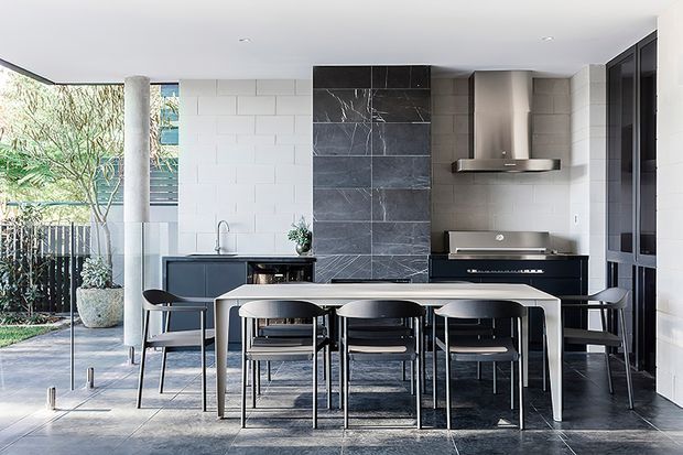A contemporary open-plan kitchen and dining area with a long white dining table and black dining chairs positioned in front of dark kitchen cabinetry. The kitchen features a dark stone-tile feature wall, pale rectangular wall tiles, a stainless-steel rangehood and cooktop, dark tiled flooring, and large glazed openings connecting to a planted outdoor area.
