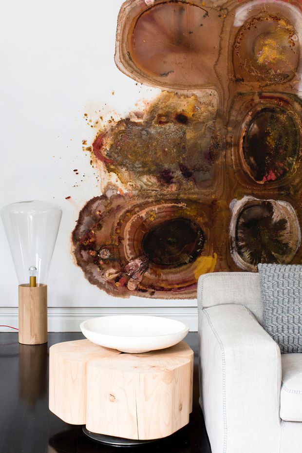 A styled living room vignette featuring a large abstract organic artwork in earthy brown, rust, charcoal and ochre tones mounted on a white wall. A pale upholstered sofa with a textured grey throw sits beside a light timber drum coffee table holding a white bowl, with a sculptural timber-and-glass floor lamp nearby. The room has a strong contrast between the dark glossy floor and light furnishings.