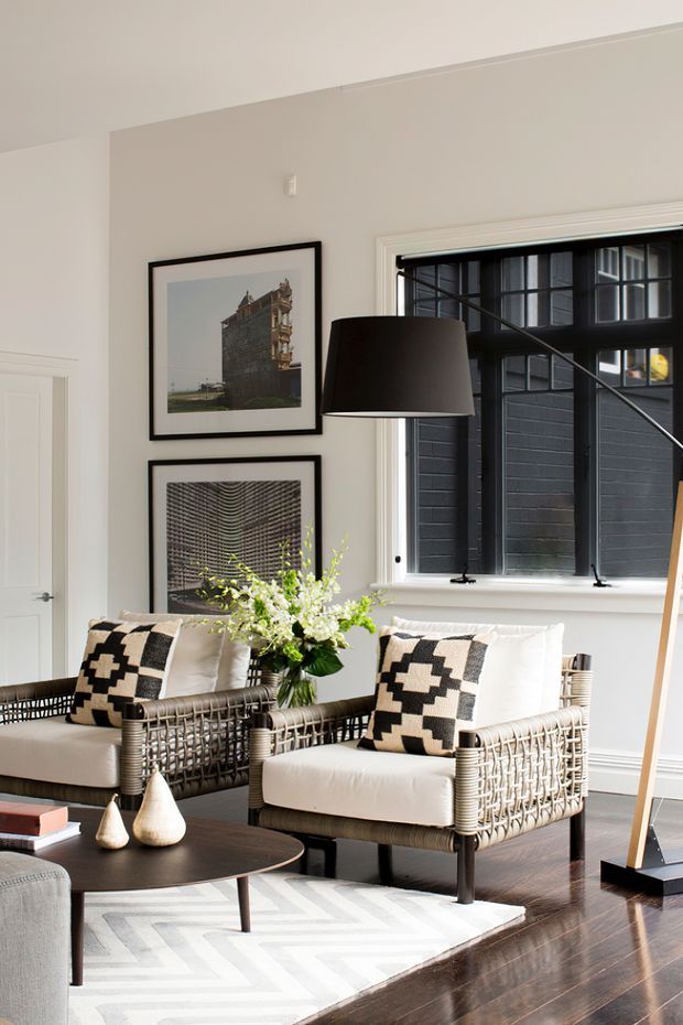 A refined living room with white walls, dark polished timber flooring, two woven armchairs with cream upholstery and geometric black-and-cream cushions, and a round dark coffee table. A large black floor lamp stands beside a gridded black-framed window, while framed architectural photographs and a simple floral arrangement add visual interest. The room has a bright, high-contrast, contemporary feel.