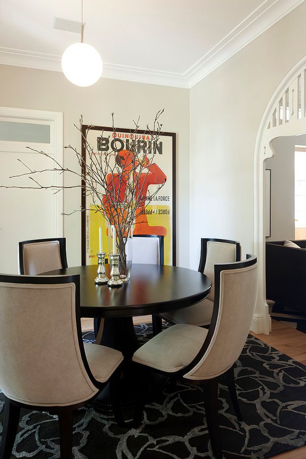 A refined dining room with a round dark timber dining table surrounded by upholstered dining chairs. The room features pale neutral walls, ornate ceiling cornices, a large framed artwork, a sculptural pendant light, patterned dark rug, timber flooring, and an arched opening leading toward an adjacent living area.