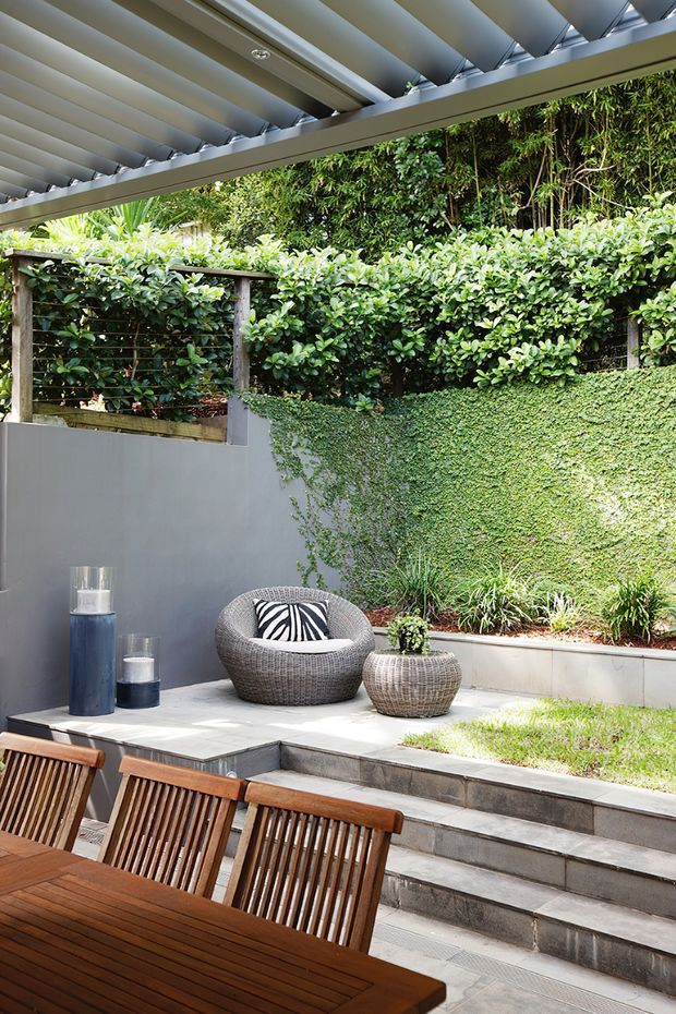 A landscaped outdoor courtyard or patio beneath a contemporary louvred roof. The space features paved stepped terraces, a rendered grey wall, dense clipped hedging and climbing greenery, timber outdoor dining furniture, woven lounge seating, and decorative planters. The setting is bright, leafy, and private, with a strong indoor-outdoor entertaining character.