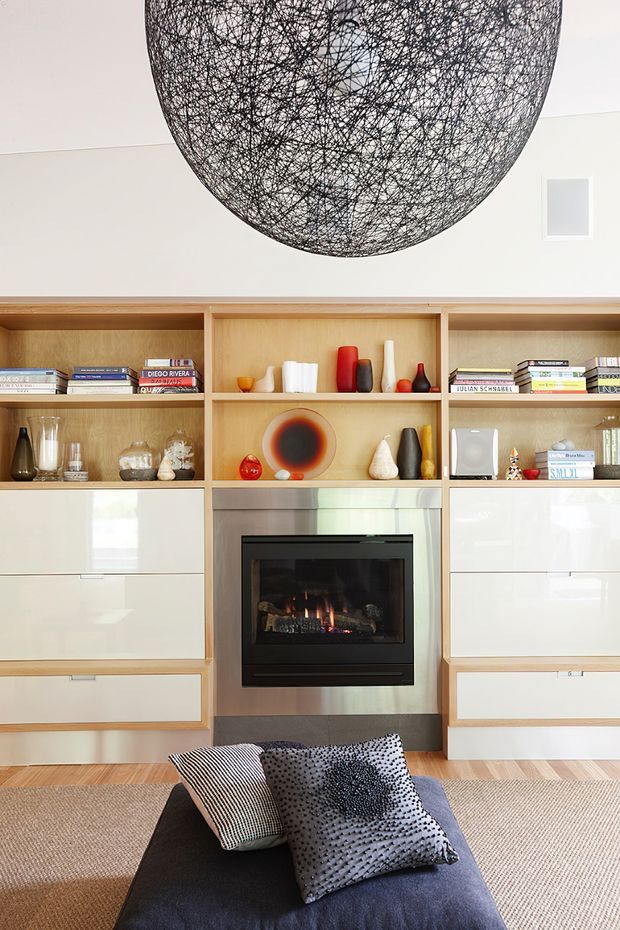 Bright contemporary living room featuring a built-in timber and glossy white storage wall with open display shelving, a central gas fireplace clad in stainless steel, and a large black wire pendant light. A dark upholstered ottoman with patterned cushions sits in the foreground over a textured neutral rug.