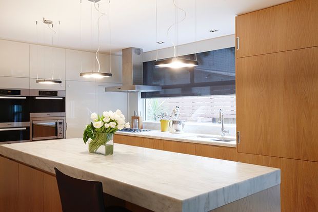 Bright contemporary kitchen featuring a large pale stone island with seating, integrated dark appliances, warm timber cabinetry, a stainless-steel rangehood, pendant lighting, and a tiled splashback behind the sink. The space has a clean, refined, highly functional fit-out with strong natural light.