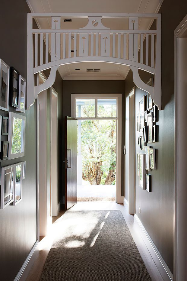 A narrow, elegant residential hallway leading to an open exterior door, with dark painted walls, white trim, carpeted flooring, framed artwork and windows along both sides. An ornate white balustrade and curved arch form a decorative overhead gallery or landing, while daylight and trees are visible beyond the doorway.