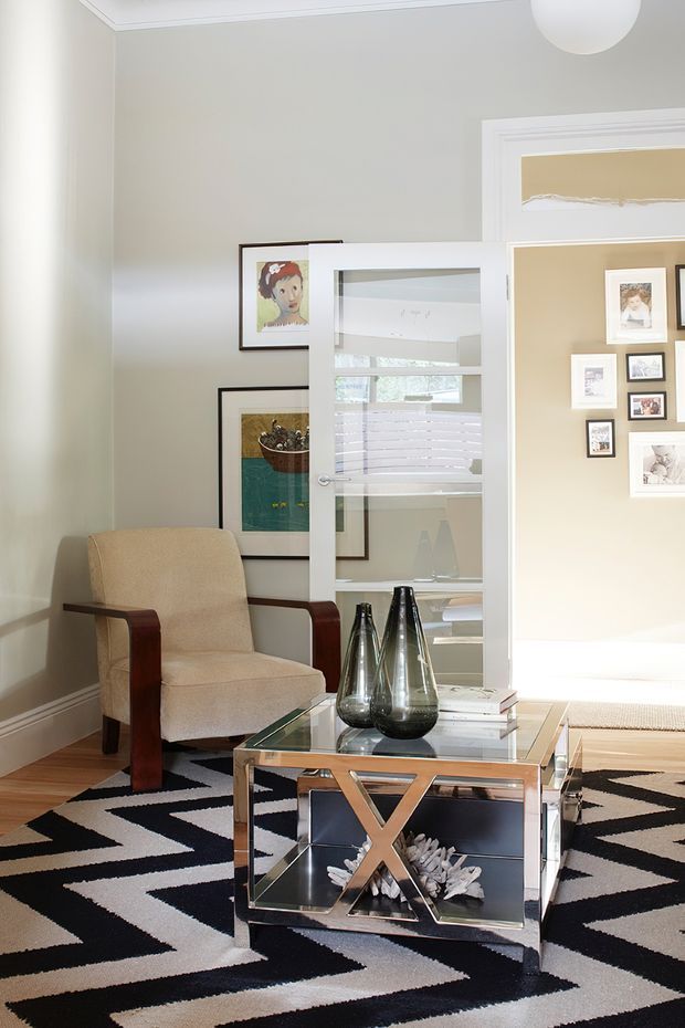 Bright, refined living room with pale grey walls, white architectural trim, timber flooring, and a bold black-and-white geometric rug. A cream upholstered armchair with dark wood arms sits beside a mirrored glass coffee table with a polished metal X-frame base. Decorative glass vases, books, framed artwork, and a glazed internal partition add layered detail. An adjoining gallery-like room is visible through the opening.