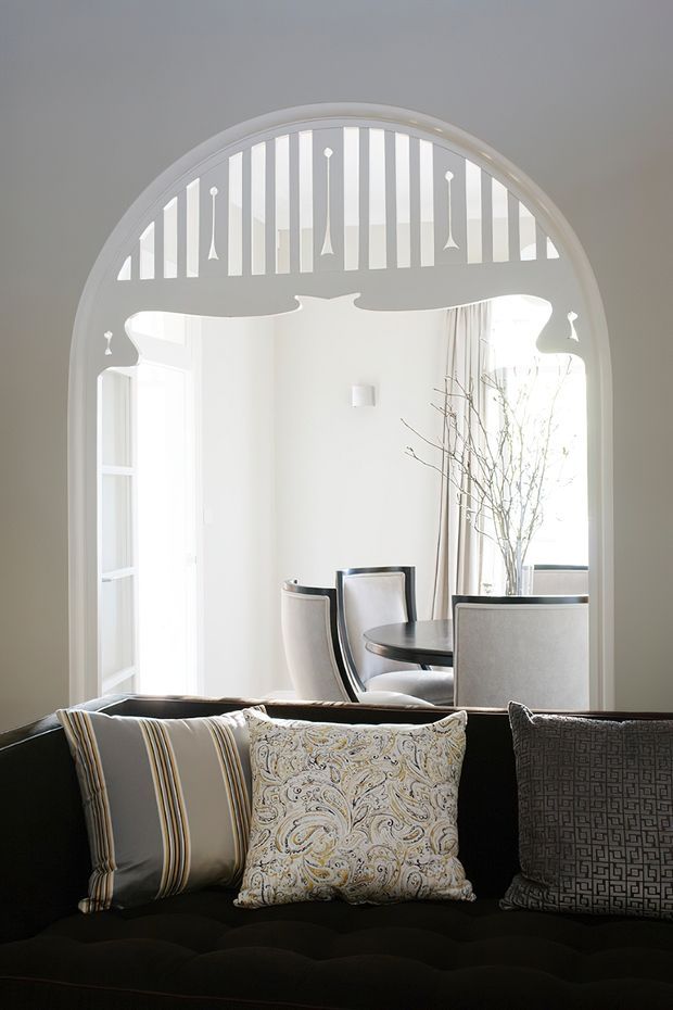Interior view framed by an ornate arched opening with decorative vertical balusters and curved detailing. A dark upholstered sofa with patterned cushions occupies the foreground, while a bright white dining area with a round dark table, upholstered dining chairs, tall curtains and a large window is visible beyond. The space has an elegant, high-contrast, traditional decorative character with abundant natural light.