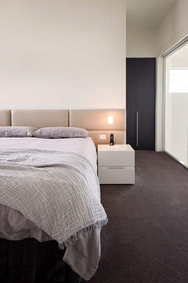 A contemporary bedroom with a neatly made bed, upholstered full-width headboard, soft neutral bedding, floating white bedside storage, and a warm wall-mounted pendant light. The room has dark carpet, a tall dark timber door, white walls, and large glazed sliding doors bringing in natural light.