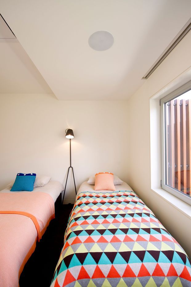 Bright compact bedroom with two single beds positioned side by side, colourful geometric bedding and coral-toned throws, white walls and ceiling, a simple black floor lamp, and a large side window with exterior vertical timber screening. The room has a clean, playful contemporary feel.