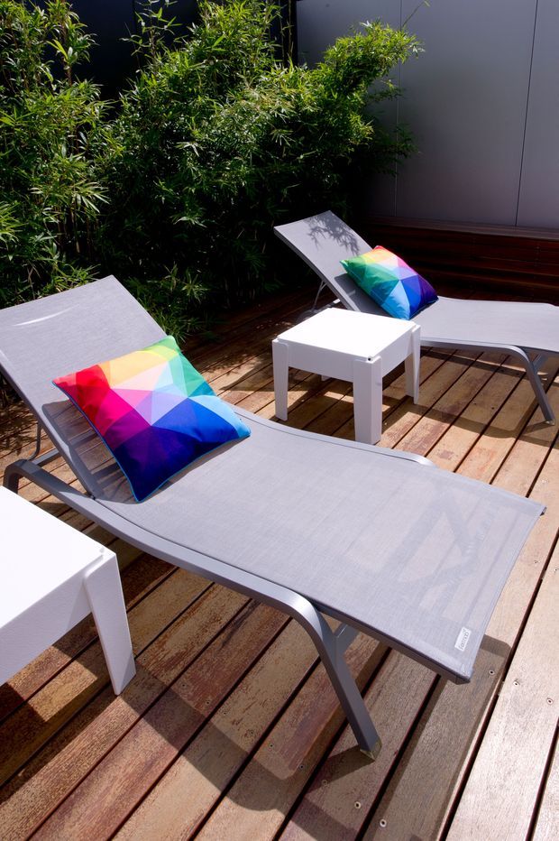 A sunny outdoor terrace furnished with multiple grey sling loungers, colourful geometric cushions, and white square side tables. The furniture sits on a warm timber deck beside dense bamboo-like planting and a dark vertical-panel wall, creating a private landscaped outdoor relaxation area.