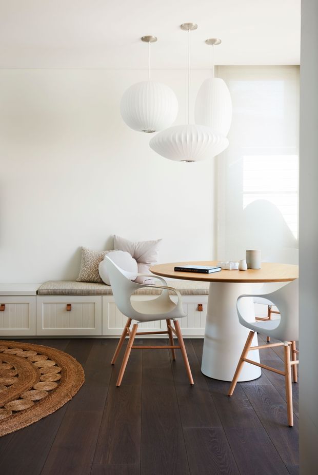 Bright, refined dining nook with a built-in upholstered window seat and white paneled cabinetry, a round wood dining table, sculptural white pedestal base, modern white chairs, and three ribbed pendant lights. Dark timber flooring and a woven round rug provide contrast against the white walls and soft filtered daylight.