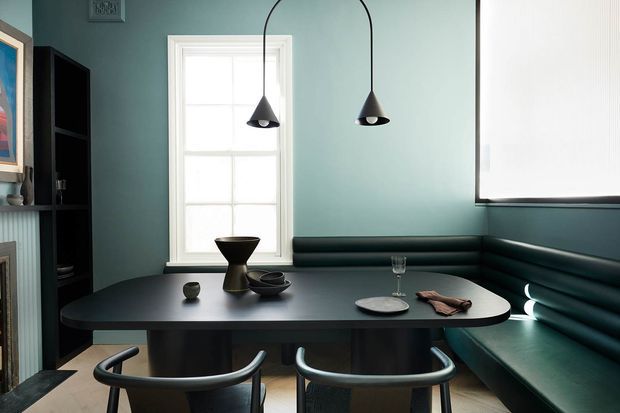 A compact, moody dining nook with a dark rounded rectangular dining table, two tubular chairs, and a built-in dark teal upholstered banquette wrapping the corner. The room has blue-green painted walls, white-trimmed windows with a roller blind, black arched pendant lights, a tall black shelving unit, and restrained sculptural tableware.