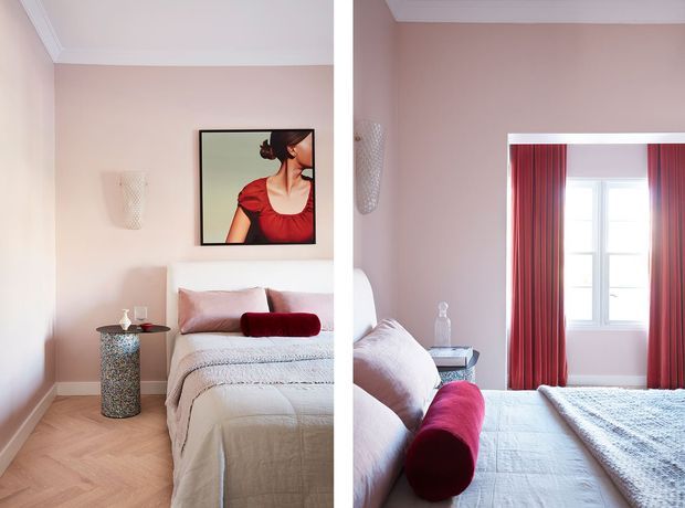 A split-view image of a refined bedroom with blush-pink walls, white decorative ceiling trim, herringbone timber flooring, and a neatly made bed dressed in pale neutral linens. The room features red velvet cylindrical bolster cushions, matching red curtains framing a bright window, a small metallic bedside table, a framed artwork, and subtle decorative lighting. The overall look is elegant, warm, and coordinated.