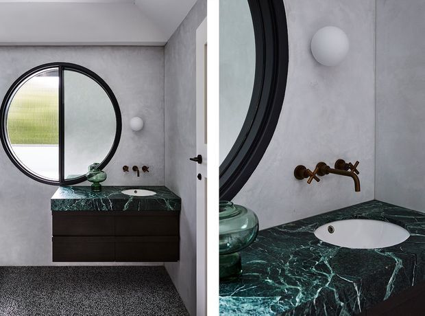 A compact, refined bathroom featuring a dark green veined stone vanity benchtop, integrated white oval basin, dark timber cabinetry, grey plastered walls, and a large round black-framed window or mirror. Brushed bronze wall-mounted tapware and a spherical wall light add warmth and contrast. The image includes a wider bathroom view and a close-up detail of the vanity.