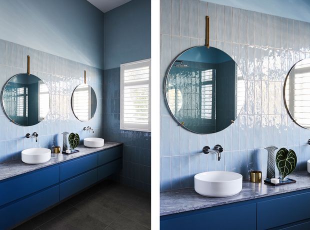 A contemporary bathroom vanity shown in a split view, featuring deep blue cabinetry, a dark stone benchtop, white vessel basins, pale blue vertically arranged glazed wall tiles, round mirrors with slim dark frames, brass wall-mounted tapware, and plantation shutters. The room has a cool, polished palette with strong geometric forms and natural light.