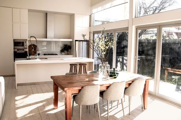 Bright open-plan kitchen and dining area with a white minimalist kitchen, large island, timber dining table, upholstered dining chairs, and extensive floor-to-ceiling glazing opening toward an outdoor garden. The space has a high ceiling, abundant natural light, and a restrained neutral palette with warm timber accents.