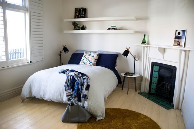 A bright, compact bedroom with a double bed dressed in white bedding and navy cushions, a patterned throw, floating display shelves, bedside lamps, and a white decorative fireplace surround with a dark inset. Pale timber floorboards, white walls, and a mustard-toned curved rug create a clean, relaxed interior.