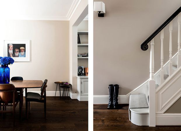 A bright, refined interior showing a dining area beside a traditional staircase. The dining space has a dark timber table and chairs, blue glass vase, framed artwork, built-in shelving, and dark timber flooring. The adjacent stair has white painted balustrades and panelling, a black handrail, carpeted treads, and a small upholstered bench beneath the stairs.