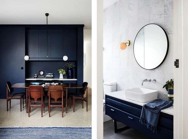 Split-view interior image showing a refined dining area with a dark navy built-in wall unit, dining table and chairs, paired with a compact bathroom featuring pale marble-look wall tiles, a round mirror, white vessel basin and navy vanity.