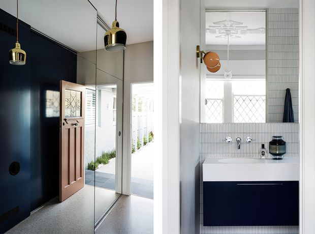 Split view of a refined residential entryway and bathroom. The entry has a dark navy feature wall, full-height mirrored panels, a timber front door with decorative glazing, and paired brass pendant lights. The bathroom features a white wall-mounted vanity with a deep navy cabinet, vertically ridged light tile walls, chrome tapware, a freestanding vessel basin, and a decorative ceiling light visible through the doorway.