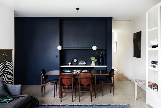 A refined open-plan dining and living area featuring a dark navy built-in cabinetry wall with integrated shelving and a long dining table surrounded by timber chairs. White walls, pale timber flooring, a textured rug, sculptural pendant lights, artwork, and open white shelving create a high-contrast contemporary interior.