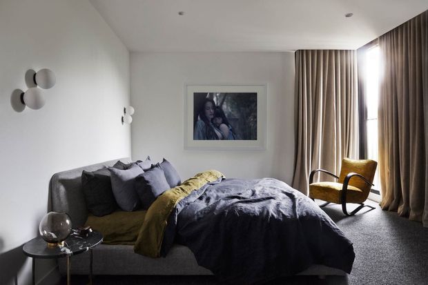 A softly lit bedroom with a grey upholstered bed, layered dark grey and muted yellow bedding, a matching grey sofa-like bed base, and a mustard yellow rocking chair. Full-height taupe curtains frame the window, while white walls, recessed ceiling lights, a framed artwork, and sculptural wall lights create a calm, refined interior.