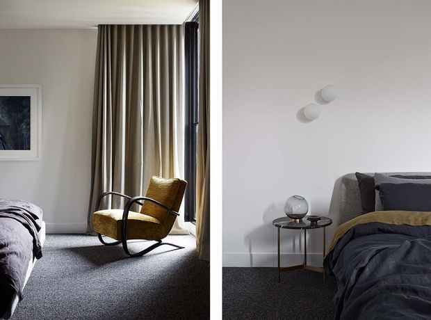 Split-view bedroom interior featuring a textured dark carpet, neutral white walls, floor-to-ceiling taupe curtains, and large windows. One view shows a mustard upholstered rocking chair beside the bed; the other shows layered dark bedding, a small round side table, globe-like decor, and minimalist wall-mounted lights. The overall design is calm, refined, and contemporary with mid-century-inspired furnishings.