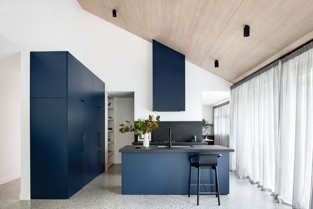 Contemporary open-plan kitchen with deep navy-blue cabinetry and island, integrated sink and tapware, terrazzo-style flooring, white walls, and a pale timber-lined sloping ceiling. Full-height sheer curtains provide soft daylight along the glazed living edge, while a single bar stool and minimal pendant-free lighting reinforce the clean, refined design.