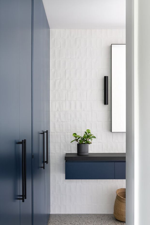 Contemporary bathroom vanity area viewed through a doorway, featuring deep navy-blue cabinetry with long black pulls, a dark benchtop, white textured rectangular wall tiles, a tall mirrored cabinet, and a terrazzo-like floor. A potted plant and woven basket add natural accents.