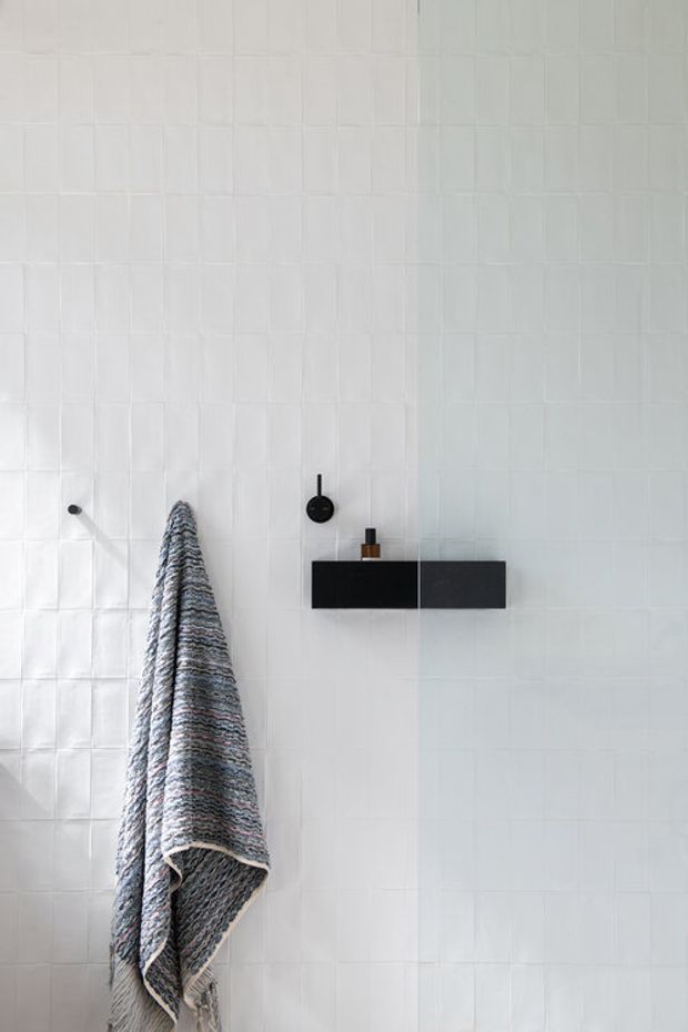 Minimalist bathroom wall clad in white textured square tiles, with a black wall-mounted floating vanity or shelf, black circular fixture, small amber container, and a hanging grey woven towel. The image focuses on clean geometric surfaces and monochromatic styling.