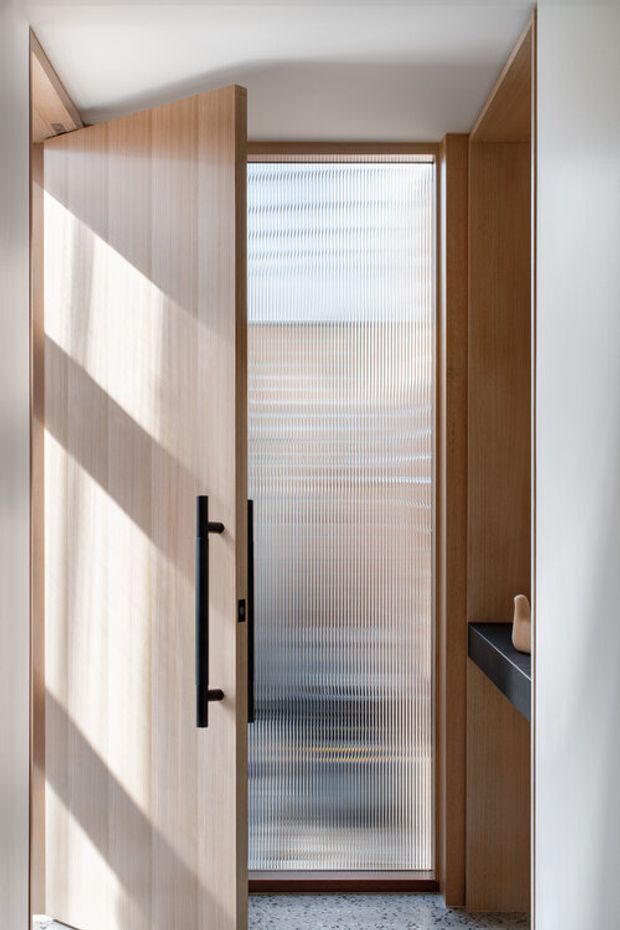 A narrow, minimalist interior passage or entry zone framed by warm vertical timber cabinetry and a large ribbed translucent glass panel. A timber door with a long black pull handle is open in the foreground, revealing a darker space beyond. A slim dark stone or composite shelf with a small sculptural object sits within the timber joinery on the right. Strong diagonal sunlight creates pronounced shadows across the pale timber door and light terrazzo-like floor.