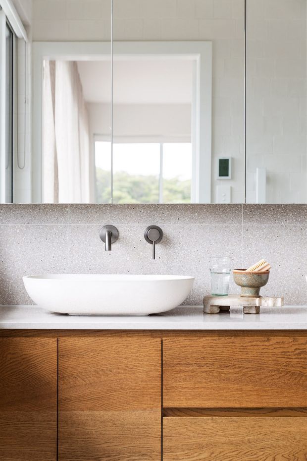 Close-up of a refined bathroom vanity with a white vessel basin, wall-mounted brushed metal tapware, a speckled grey terrazzo-like tiled backsplash, and warm timber cabinetry with integrated drawers. A large mirror reflects a bright adjoining space and window, while small toiletries sit on the benchtop.